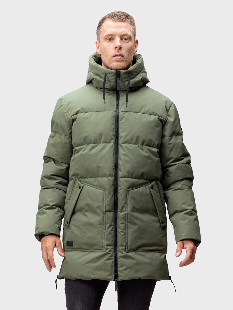 En man bär Halti Linjat Parka Jacka Herr, en lång grön puffer med huva, vänd framåt mot en grå bakgrund.