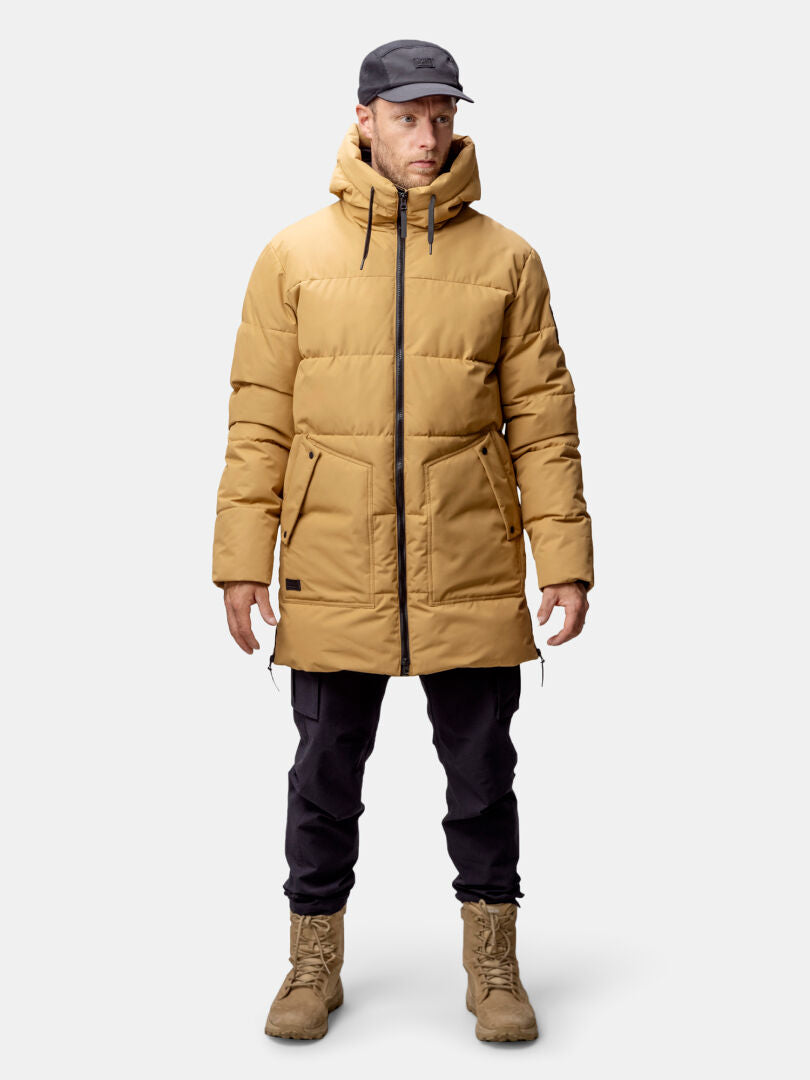 En man i brun Halti Linjat Parka Jacka Herr, svarta byxor och stövlar står vänd framåt mot en enfärgad bakgrund.