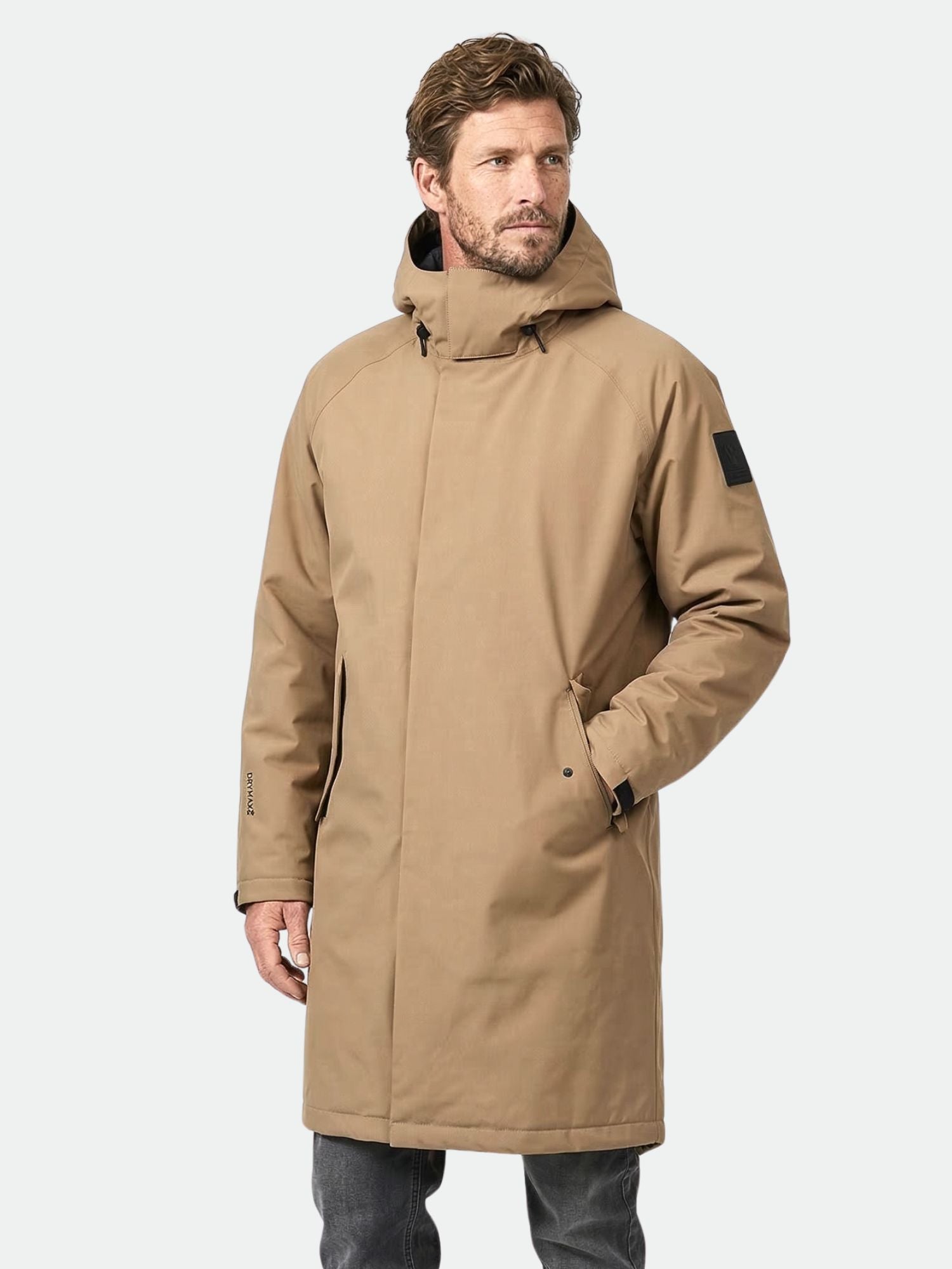 En man bär Halti Flemari DX Parka Jacka Unisex i beige och står med ena handen i fickan mot en enfärgad bakgrund.