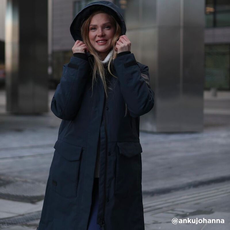 En person i en Halti Bergga Dam DrymaxX Vinterjacka står utomhus och håller i huvan med båda händerna.
