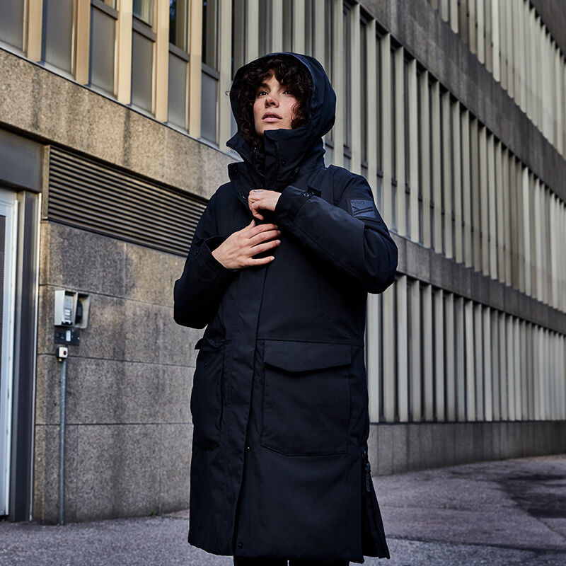 En person i en svart Halti Bergga DX Vinterjacka Dam står utomhus vid en modern byggnad med stenväggar och fönster.