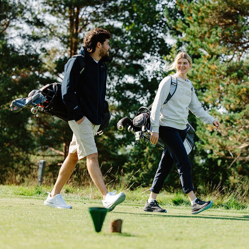 Två personer går på en golfbana i Halti Drive X-stretch Chinos Dam, med träd bakom och grönt gräs under fötterna.
