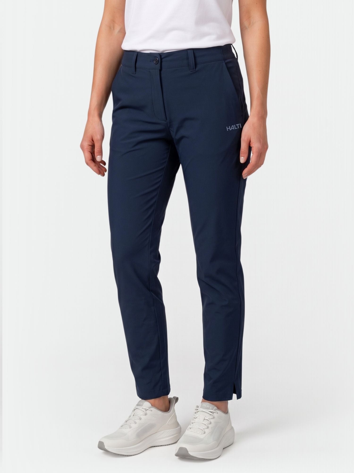 Person klädd i marinblå Halti Drive X-stretch Chinos Dam, vit t-shirt och sneakers, stående mot en vit bakgrund.