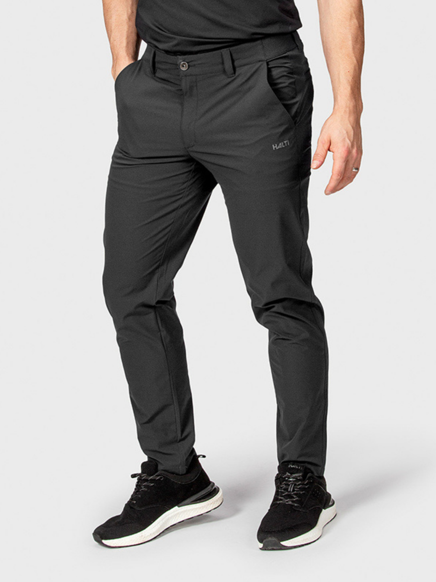 En person står med händerna i fickorna, iklädd Halti DriveX-stretch chinos herr och sportskor mot en enfärgad bakgrund.