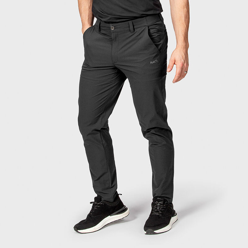En person i Halti Drive X-stretch Chinos Herr och sneakers står med händerna i fickorna mot en ljus bakgrund.