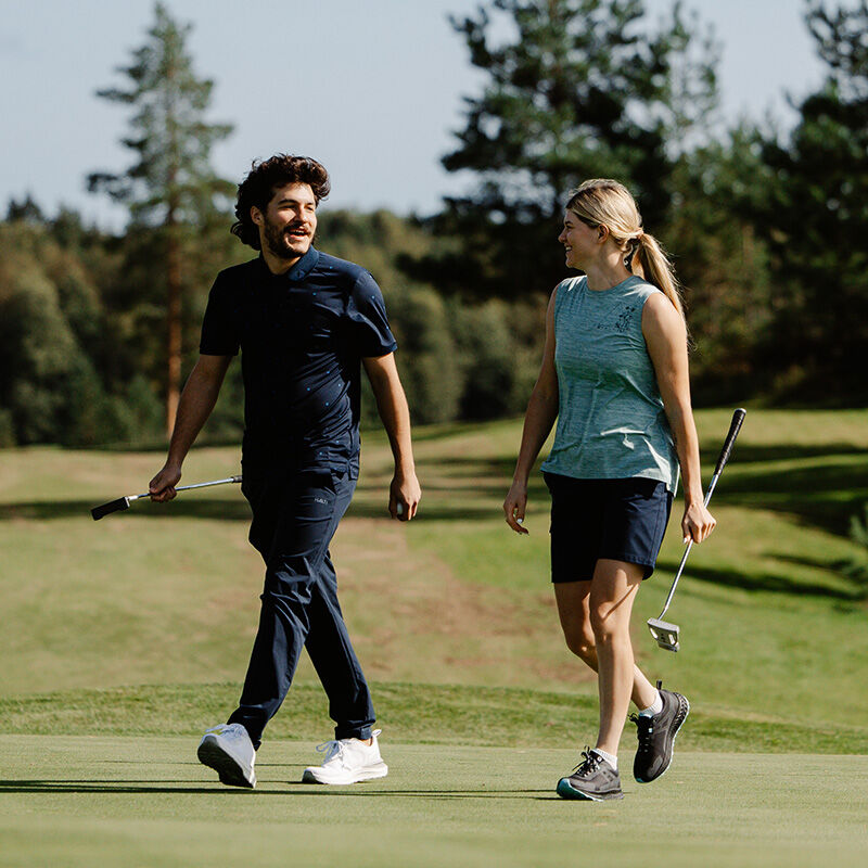 En man och en kvinna går på en golfbana i Halti DriveX-stretch Chinos Herr, var och en med varsin klubba, med träd i bakgrunden.