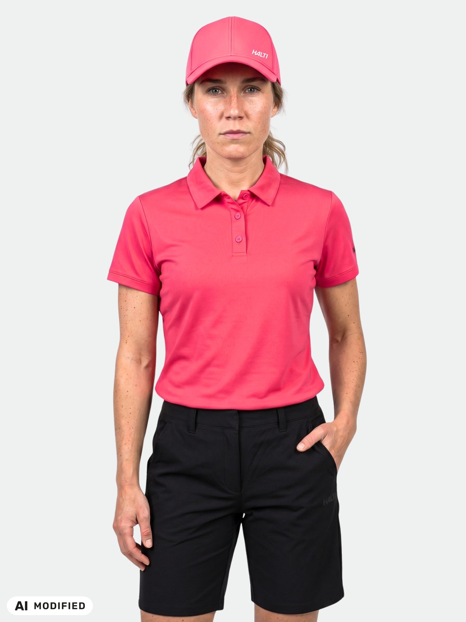 Hon bär en Halti Laine Teknisk Pikétröja för damer i rosa och vänder sig mot kameran med ena handen i fickan.