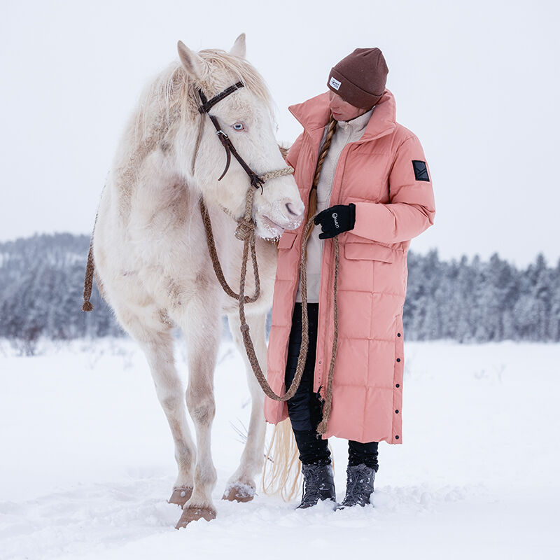 Kvinna i en lång rosa Halti Penger Vinterjacka Dam och brun hatt står bredvid en vit häst i snön.