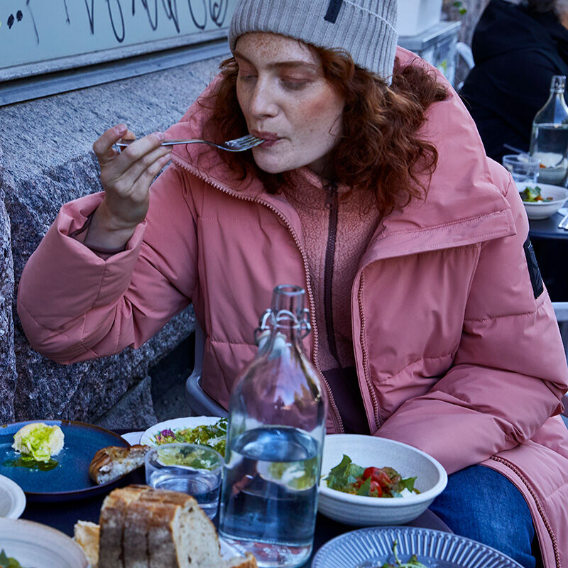 En person i en rosa Halti Penger Vinterjacka Dam och grå mössa äter utomhus med mat och vatten.