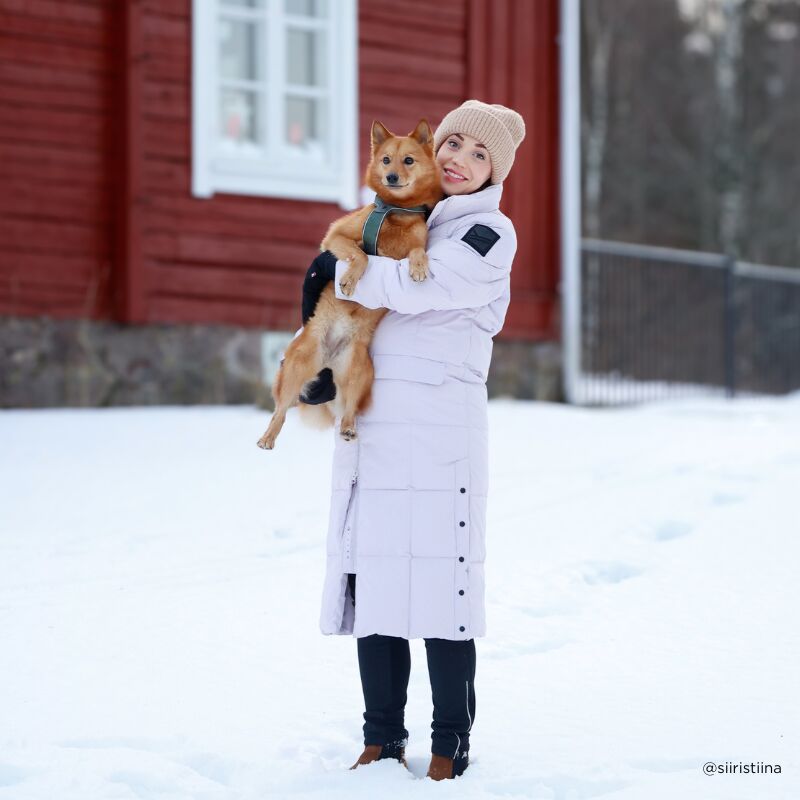 En person i en Halti Penger Vinterjacka Dam och mössa håller en liten brun hund på snö framför en röd byggnad.