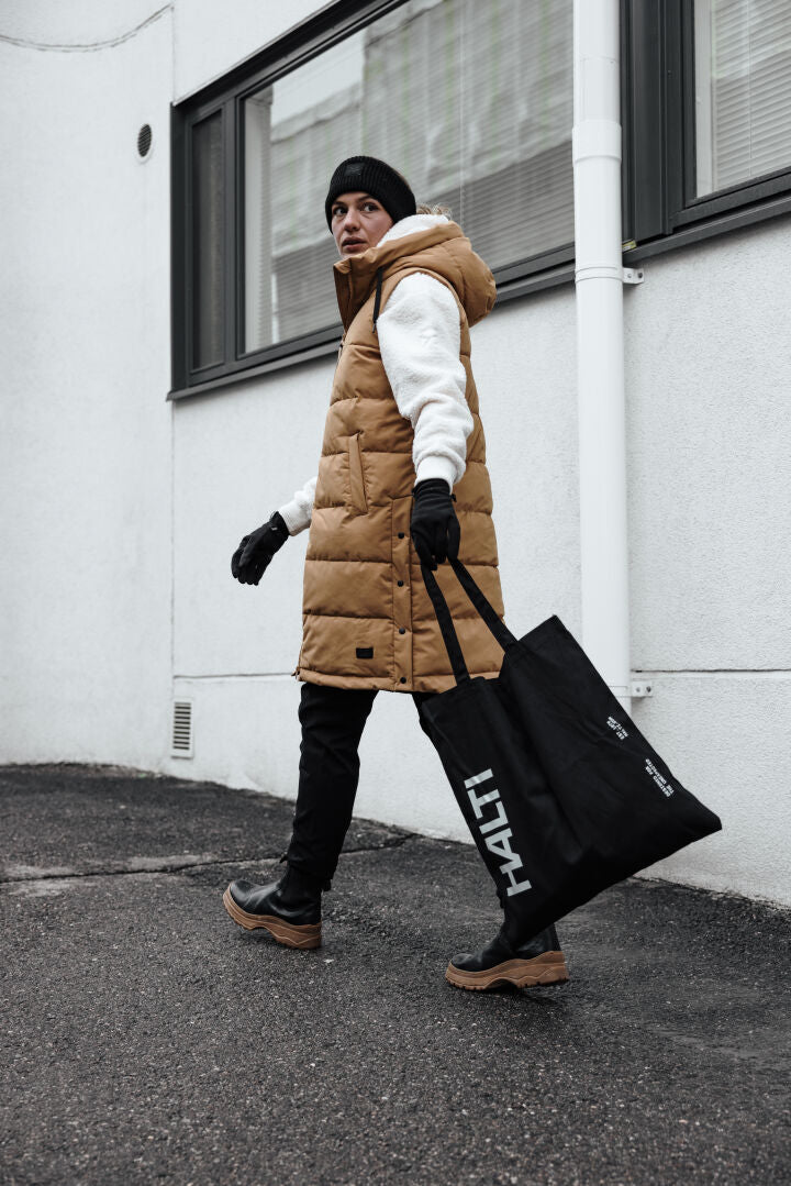 En person i brun pufferväst, svart mössa och stövlar bär en stor Halti-väska och har på sig Lykky Fleecetröjä Dam.