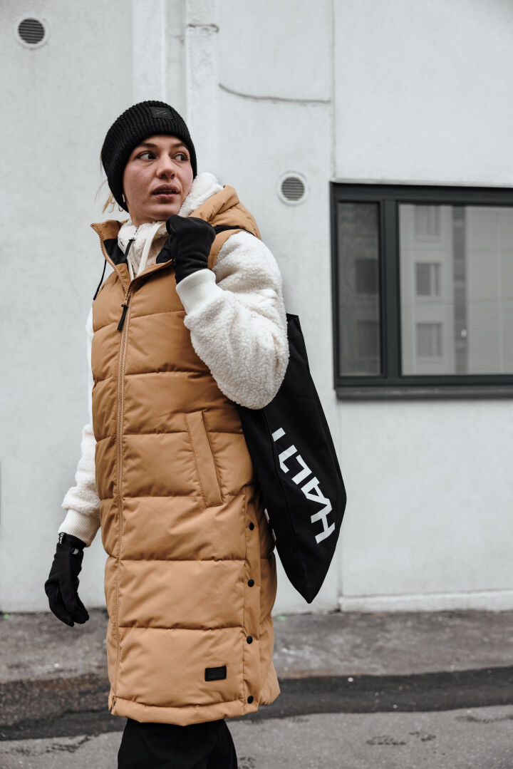 En person i brun pufferväst, Halti Lykky Fleecetröjä Dam-tröja, svart mössa och med en svart tygväska går ut.