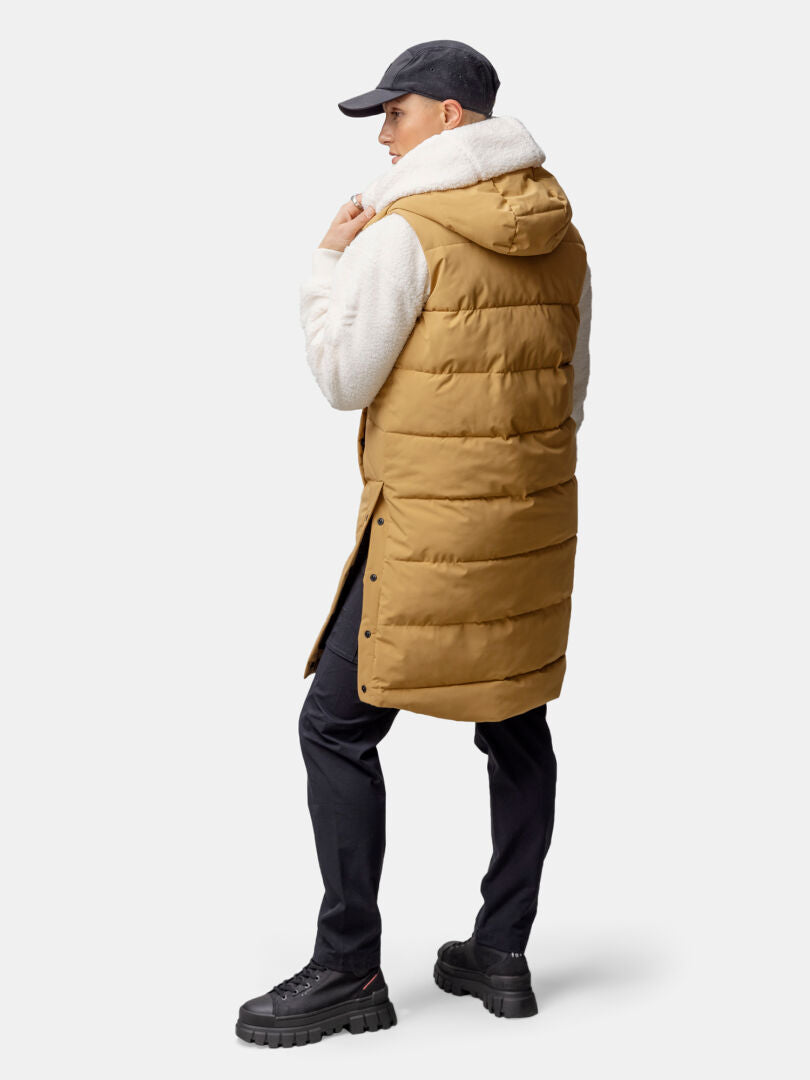 En person iklädd Haltis Penger Puffer Väst Dam i tan (återvunnen polyester) står med ansiktet vänt bort i vit huvtröja och svarta byxor.