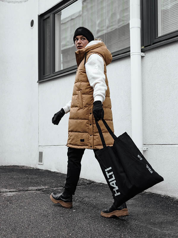 Person i brun Halti Penger Puffer Väst, vit huvtröja och svart mössa går ut med en stor svart Halti-väska.