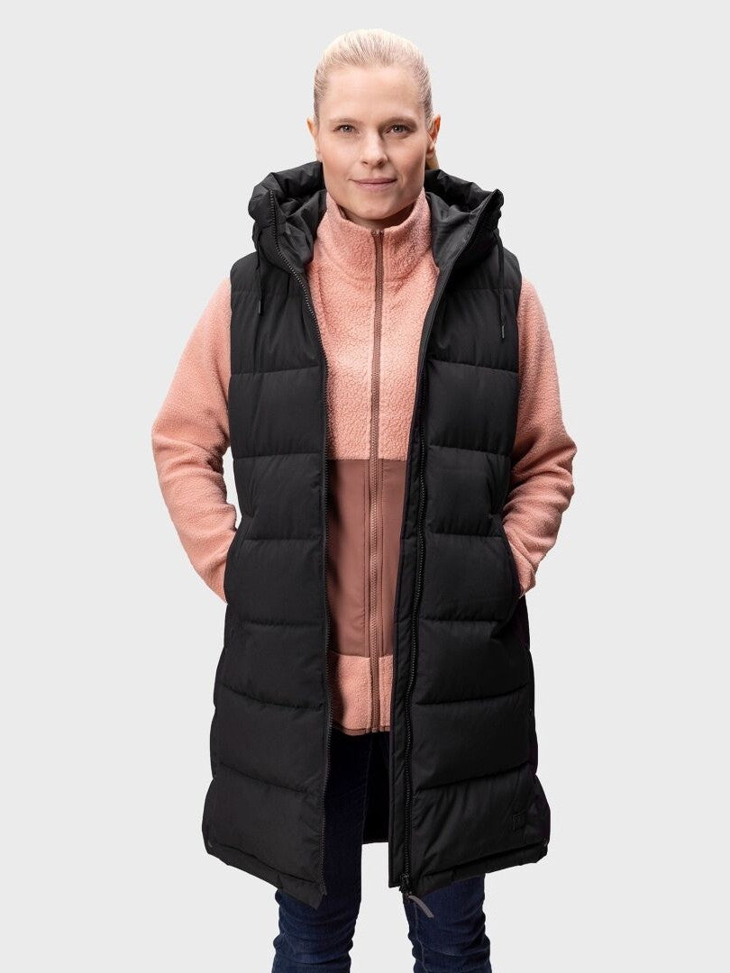 En person bär Halti Penger Puffer Väst Dam över en rosa dragkedja, med händerna i västfickorna mot en enfärgad bakgrund.