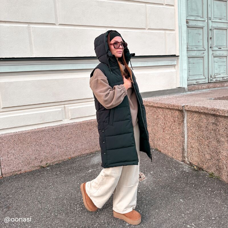 En kvinna bär Halti Penger Puffer Väst Dam, beige tröja, vida byxor och stövlar vid en stenmur.