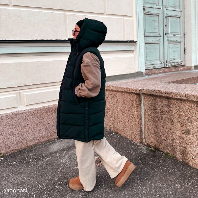 En person klädd i Halti Penger Puffer Väst Dam, beige byxor, bruna skor och tröja går förbi en ljus byggnad.