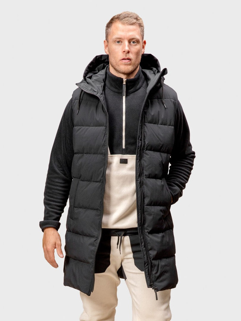 En man bär en Halti Penger Puffer Väst Herr över en svart-beige zip-tröja och beige byxor mot neutral bakgrund.