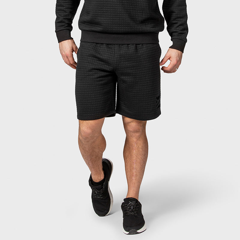 En man i en svart tröja, Halti Gliffa Shorts Unisex och svarta sneakers står mot en grå bakgrund.