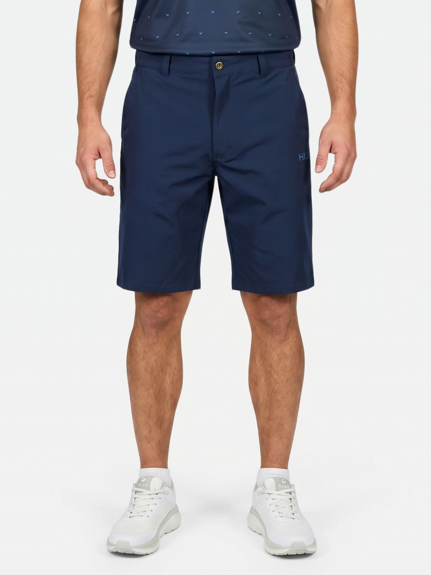 En person klädd i Halti Drive X-stretch Shorts Herr, en mörk tröja och vita sneakers står framför en enfärgad bakgrund.