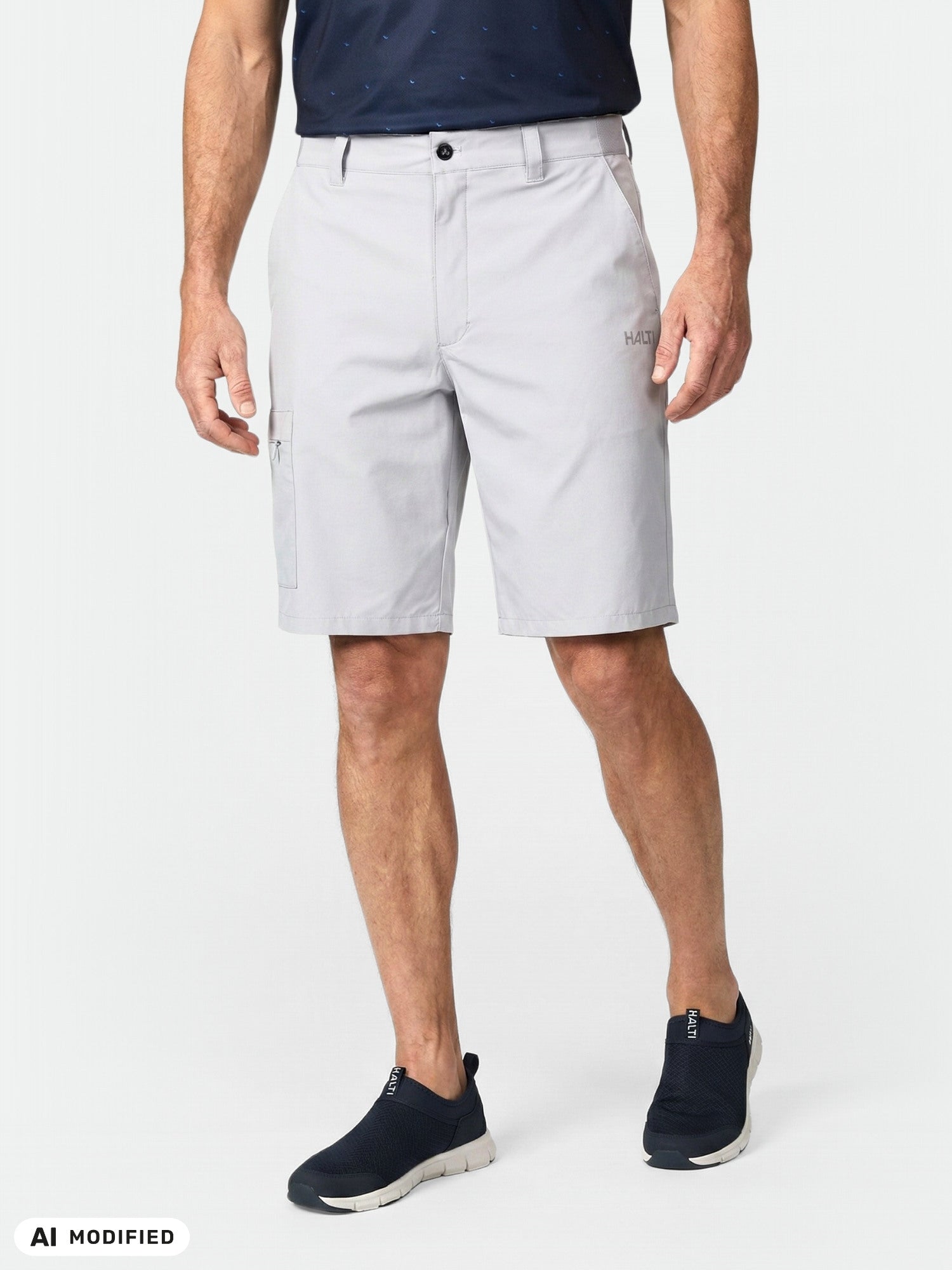 En man klädd i Halti Drive X-stretch Shorts Herr i ljusgrått, en marinblå skjorta och svarta slip-ons, framför en enfärgad bakgrund.