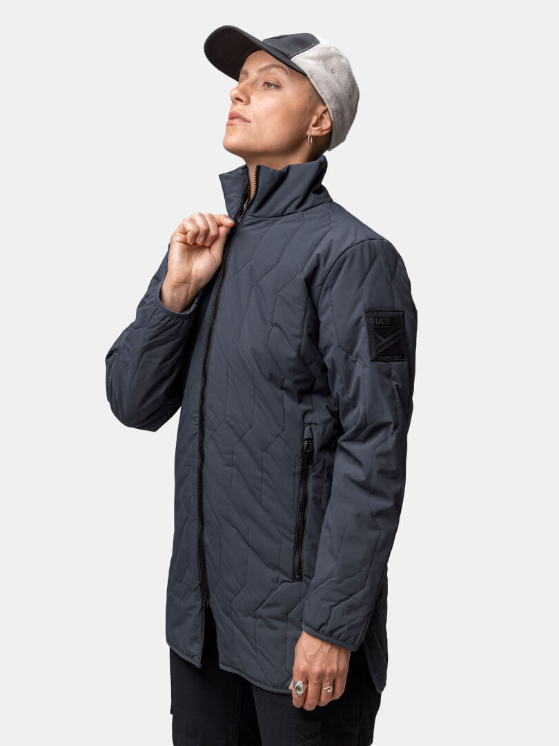 Person i Halti Synkas 3in1 DrymaxX Parka Dam och keps står mot enkel bakgrund, håller jackans dragkedja vid hakan.