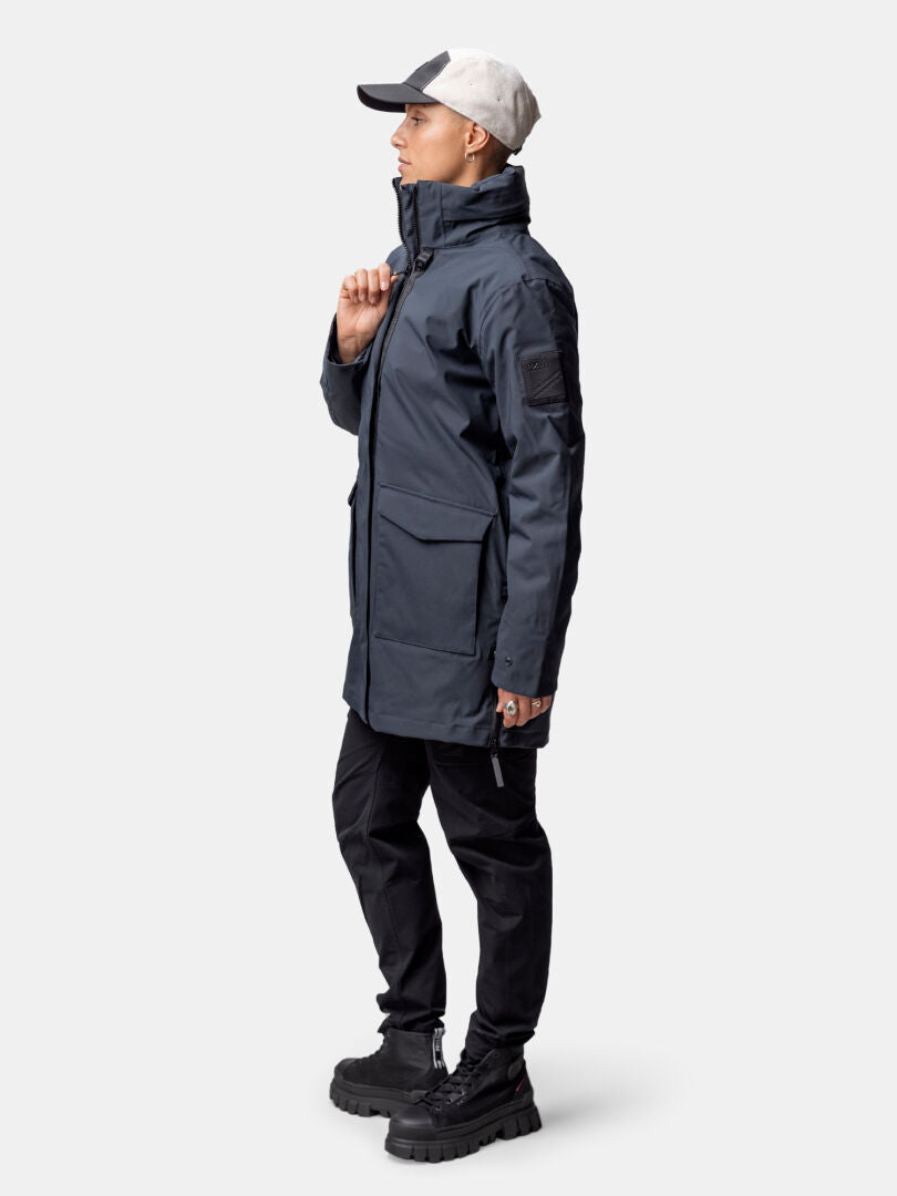 En kvinna i Halti Synkas 3in1 DrymaxX Parka står i profil, tillsammans med svarta byxor, stövlar och en grå mössa.