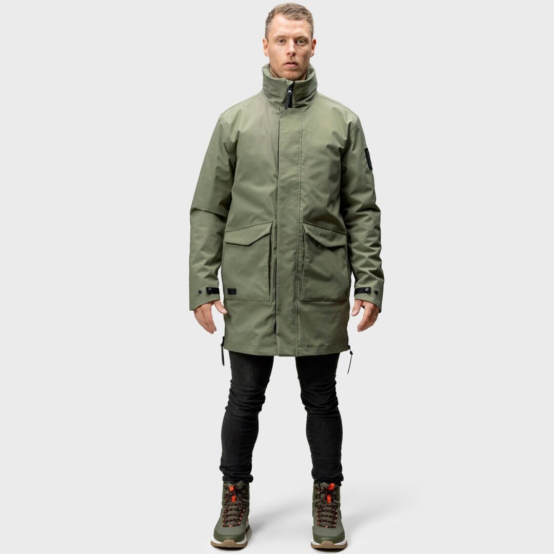 En man tittar framåt i en Halti Synkas 3in1 DrymaxX Parka Herr, svarta byxor och vandringskängor mot en enfärgad bakgrund.