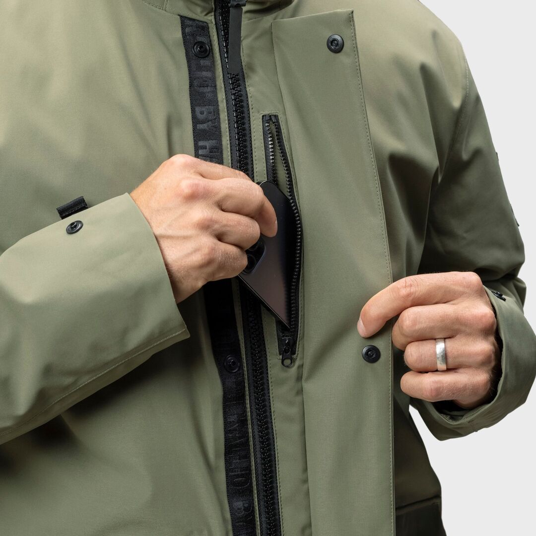 En person placerar en smartphone i den dragkedjeförsedda bröstfickan på en olivgrön Halti Synkas 3in1 DrymaxX Parka Herr.
