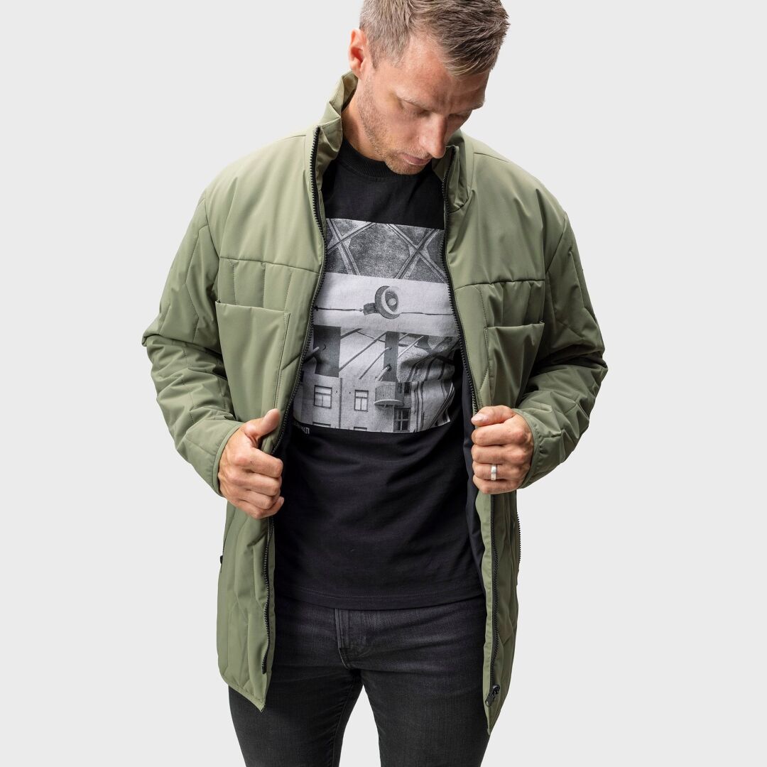 En man i en olivgrön Halti Synkas 3in1 DrymaxX Parka Herr över en svart t-shirt med mönster tittar ner och håller den öppen.