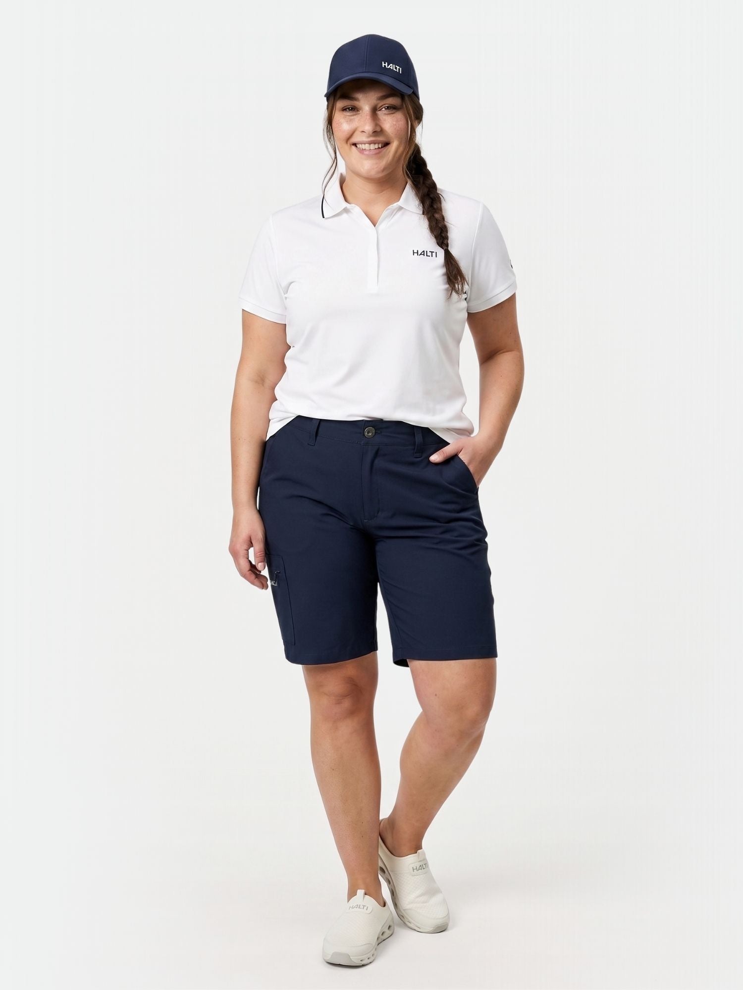 En kvinna i vit polo och Halti Drive X-stretch Shorts Dam Plus står och ler med ena handen i fickan.