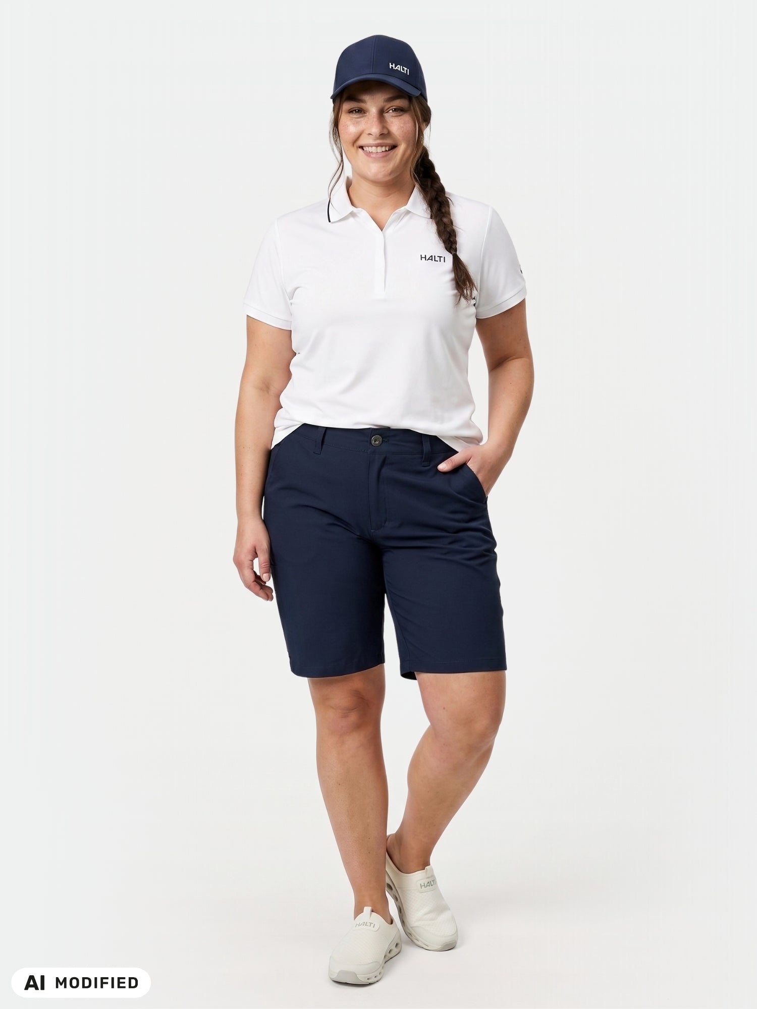 En kvinna bär Halti Drive X-stretch Shorts Dam Plus, en vit polotröja, en marinblå keps och vita skor, och ler framför en enfärgad bakgrund.