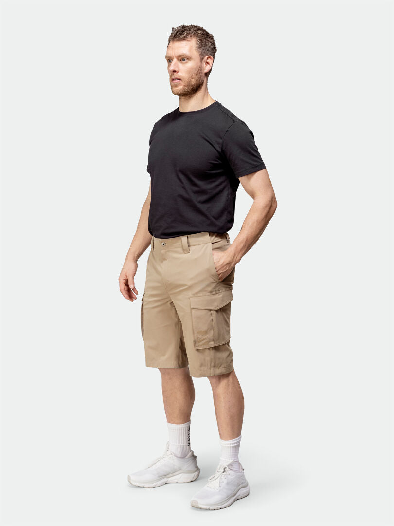 En man bär en svart t-shirt, Halti Roams Shorts Herr, vita strumpor och sneakers mot en enfärgad bakgrund.