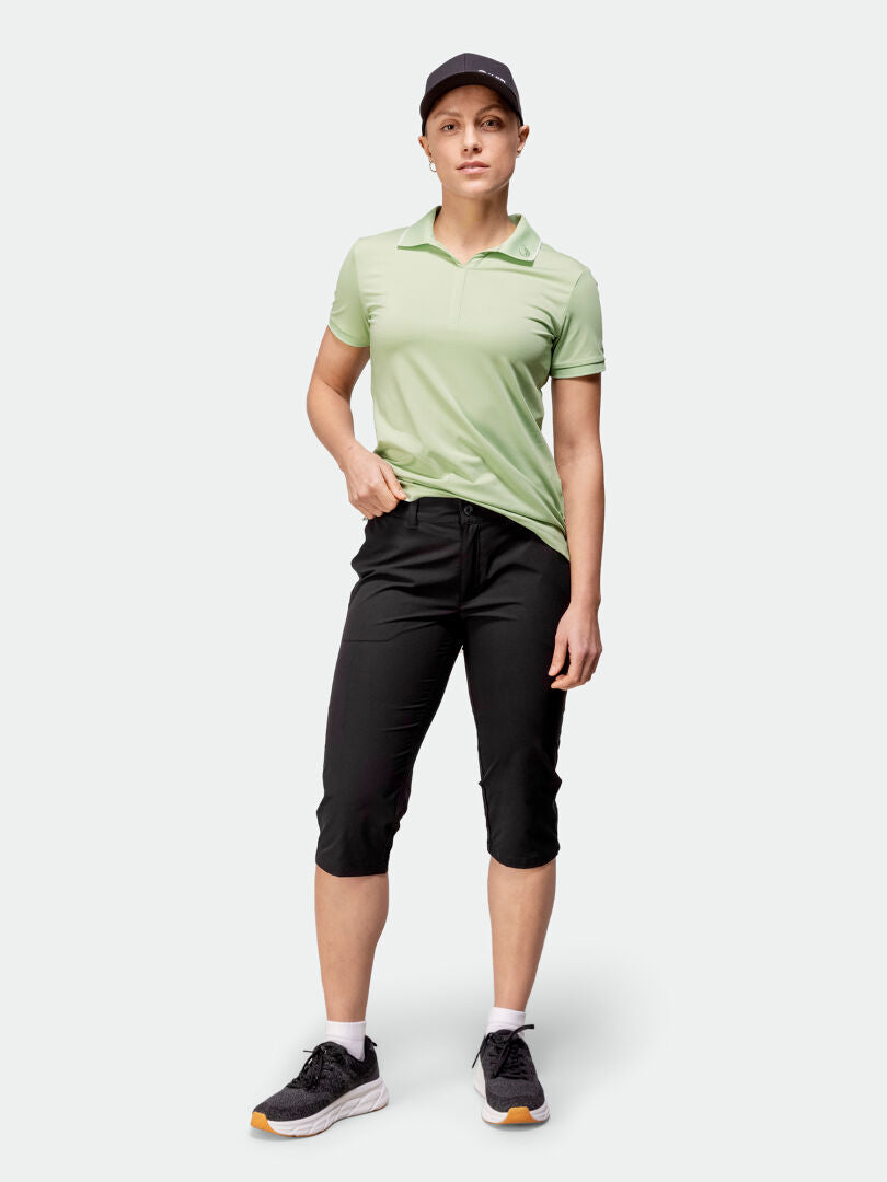 En person i grön polo, Halti Drive X-stretch Capribyxor Dam, svart keps och sneakers står vänd framåt.