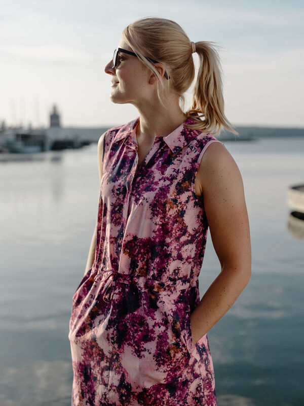 En kvinna i Halti Chipper X-stretch Klänning Dam av snabbtorkande material står vid vattnet med båtar bakom.