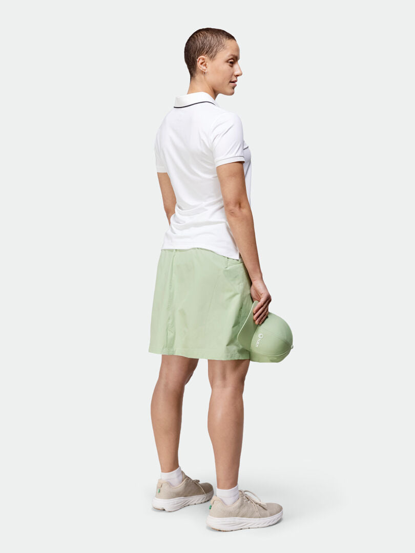 Personen står baklänges i en vit pikétröja, ljusgrön kjol från Halti Drive X-stretch Kjol Dam, sneakers och håller i en hjälm.