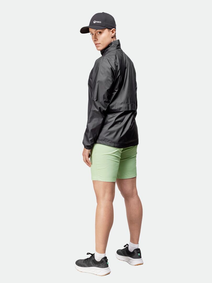 Person i Halti Saime Vindjacka Dam, gröna shorts, svart keps och sneakers står och tittar över axeln.