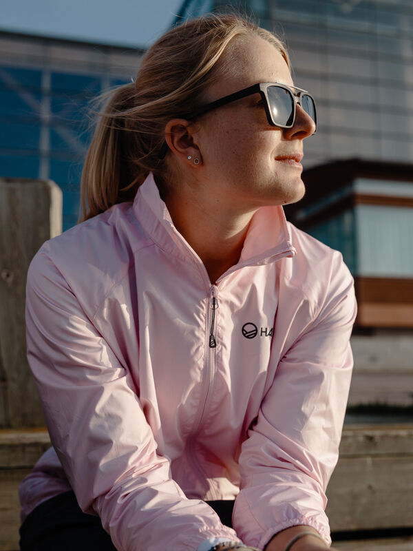 En kvinna i Halti Saime Vindjacka Dam och solglasögon sitter utomhus och tittar åt sidan i solljus.