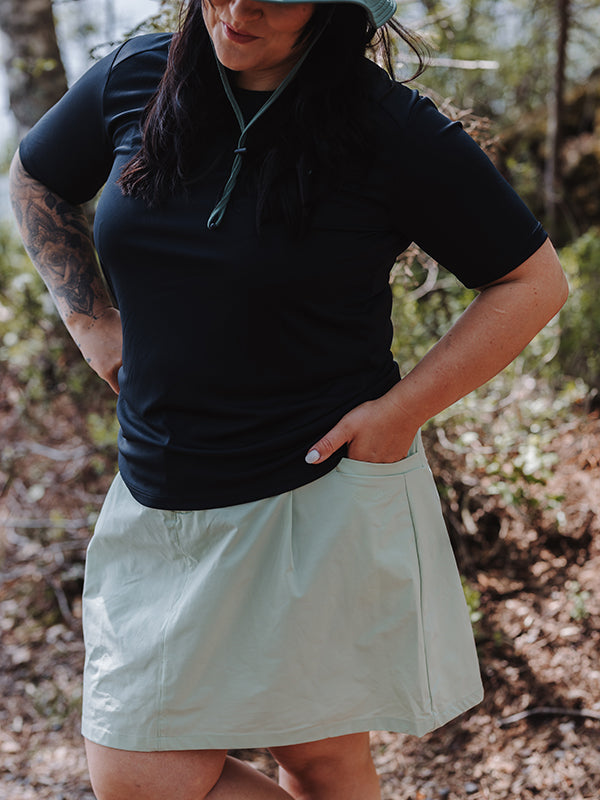 Ute i Halti Drive X-stretch Kjol Dam Plus och solhatt, händer i fickorna—perfekt för utomhusaktiviteter.