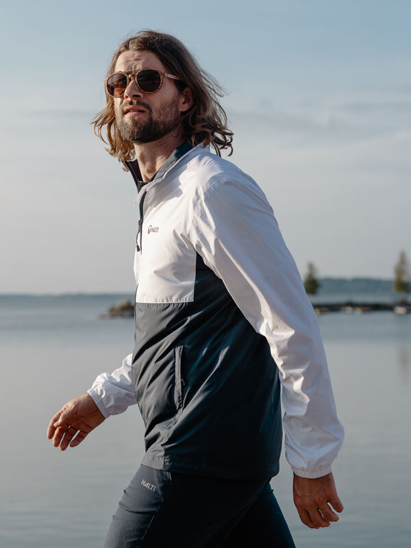 En man med långt hår och solglasögon går vid vattnet, iklädd Halti Saime Vindjacka Herr tillverkad av återvunnet material.