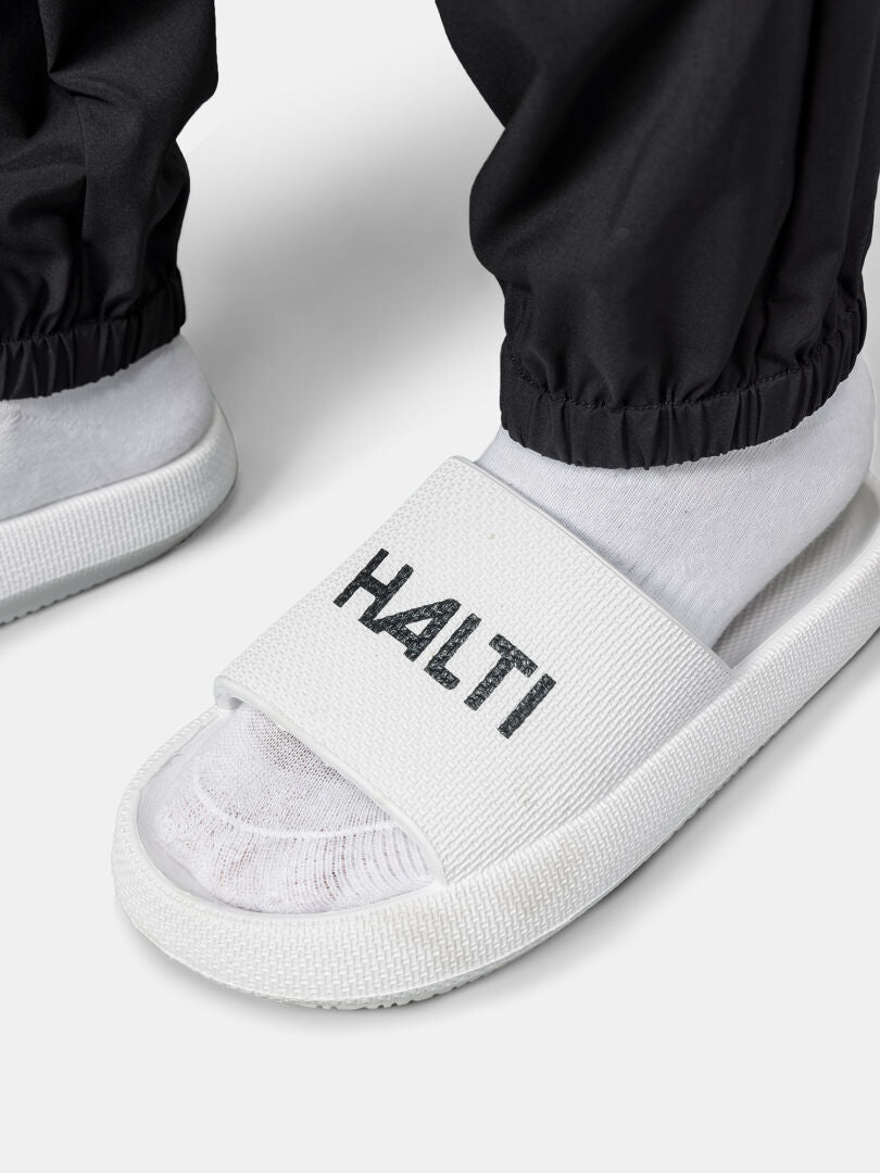 En person i svarta Halti Grets Joggers Dam och vita Halti slide-sandaler står på en ljus yta.