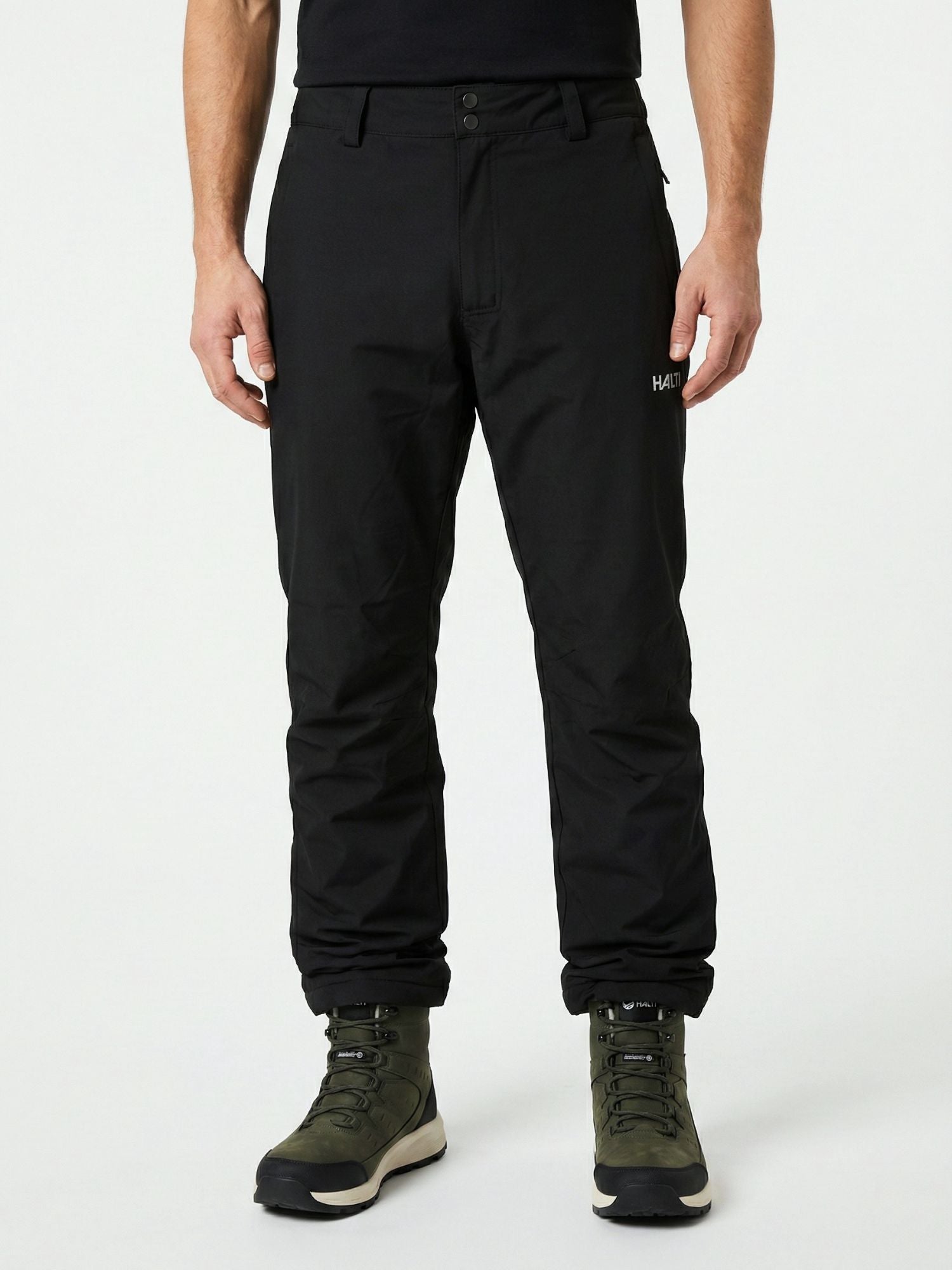 En person i svarta Halti Routa Vinterbyxor Herr-byxor, gröna vandringskängor och en svart skjorta står mot en ljus bakgrund.