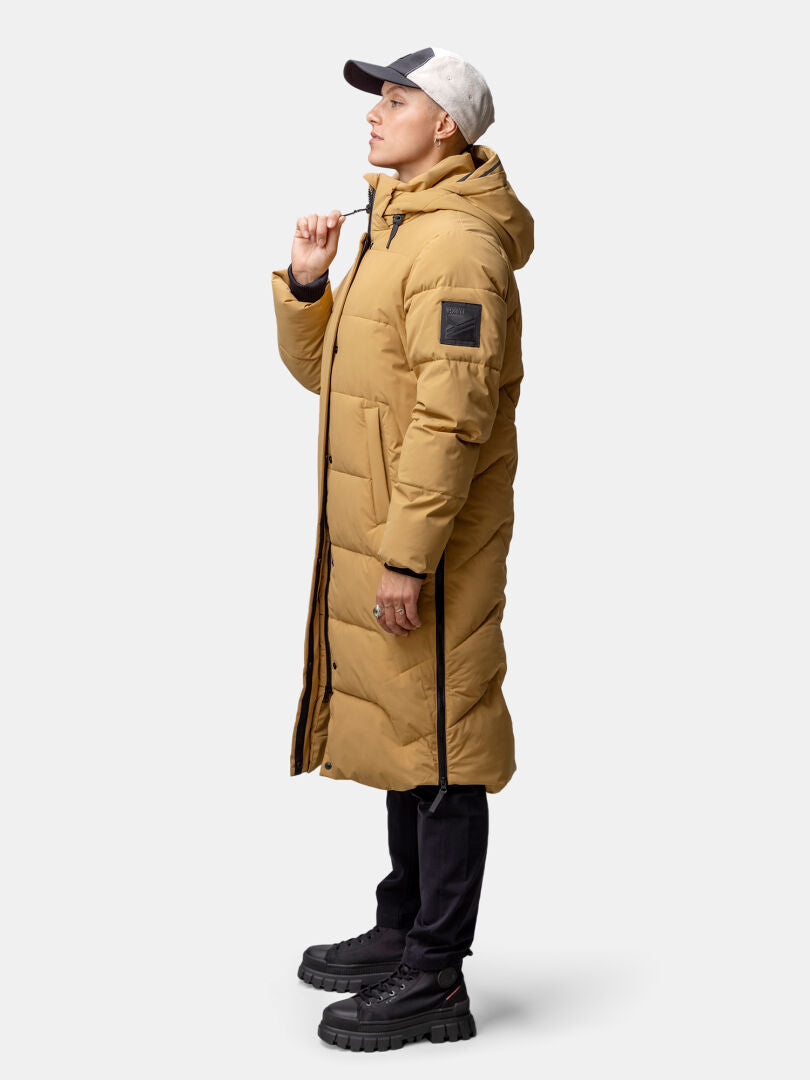 En person klädd i Haltis Brahe Puffer Long Vinterjacka Dam, svarta byxor och grå mössa står vänd mot vänster.