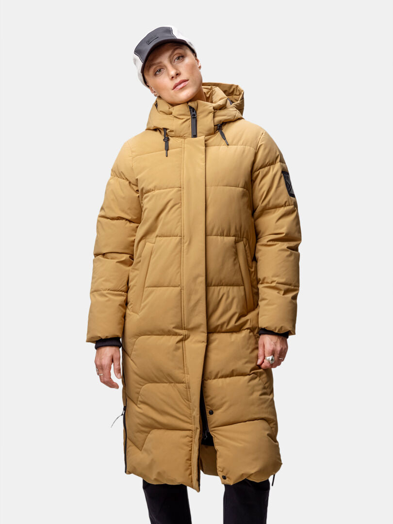 En person klädd i Haltis Brahe Puffer Long Vinterjacka Dam, svarta byxor och keps står mot en enfärgad bakgrund.