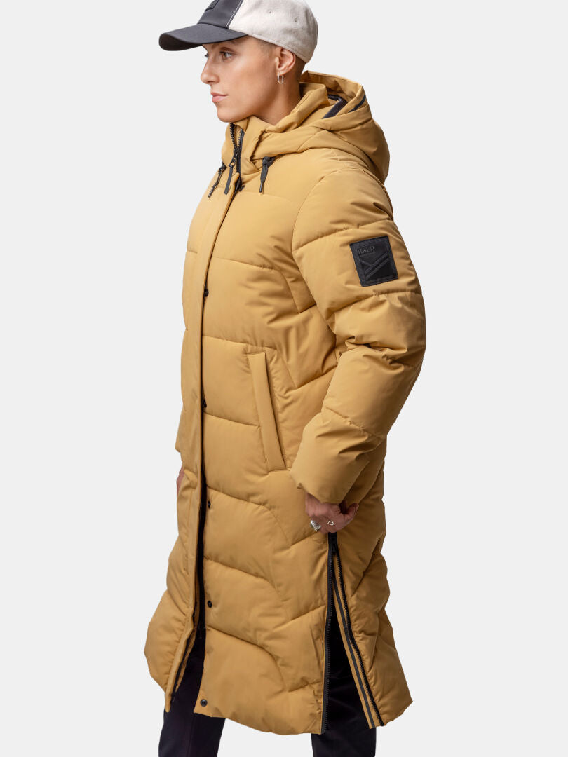 Modell i Halti Brahe Puffer Long Vinterjacka Dam, svarta byxor och ljus keps mot vit bakgrund.