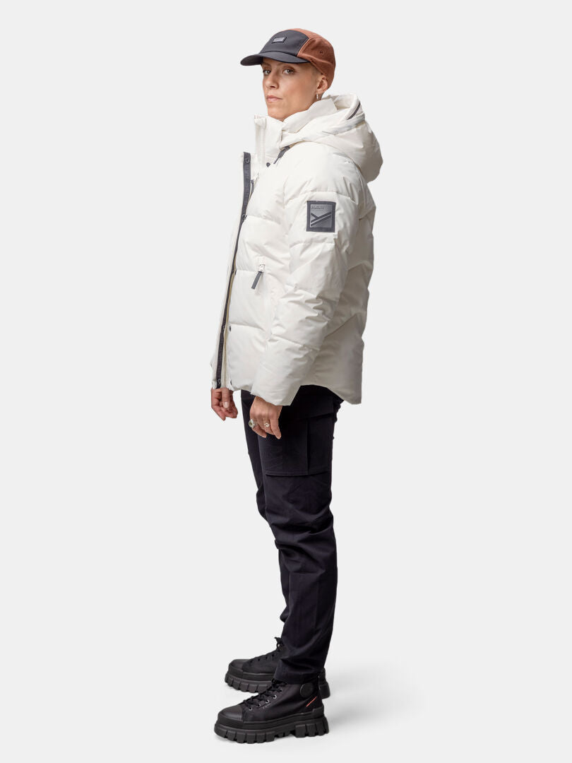 Person stående i sidled i vit Halti Brahe Puffer Vinterjacka Dam, svarta byxor och tvåfärgad keps.
