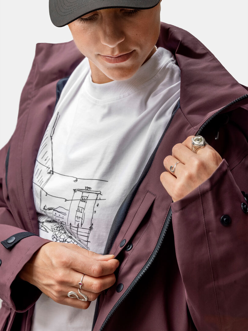 Person klädd i en rödbrun Halti Tokoi II DX Parkajacka Dam, med dragkedja, över en grafisk t-shirt, med ringar på fingrarna.
