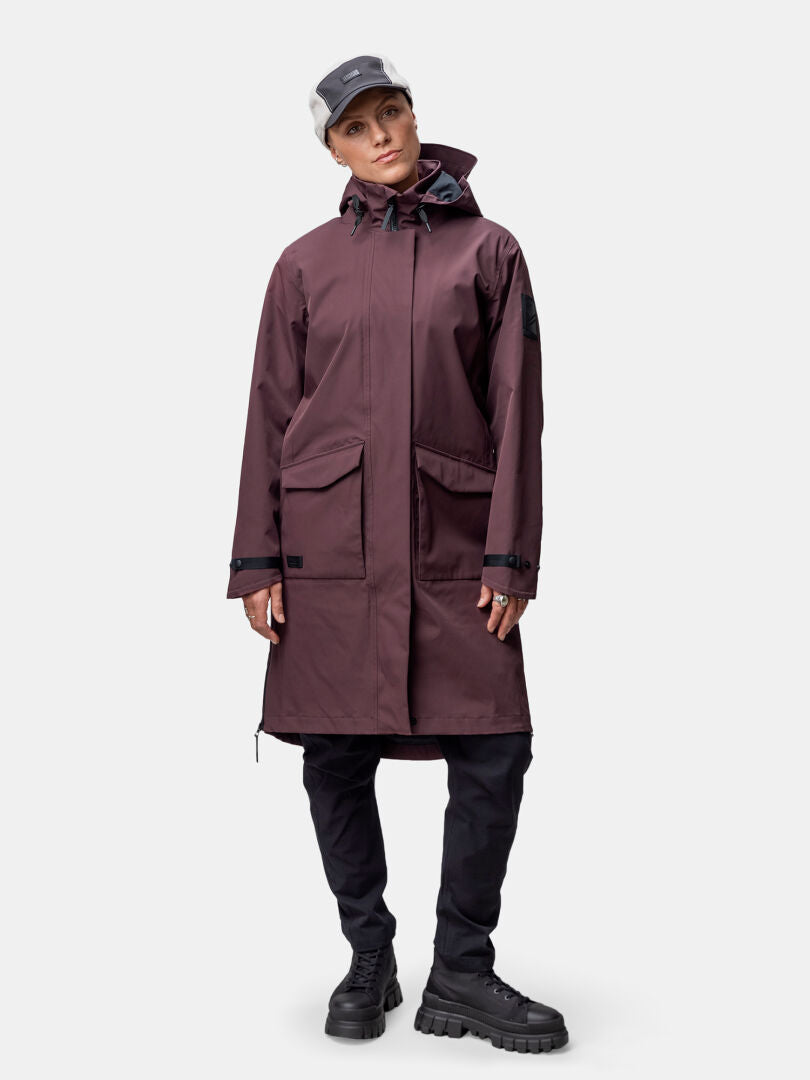 En person bär en rödbrun Halti Tokoi II DX Parkajacka Dam, svarta byxor, stövlar och en grå mössa mot en enfärgad bakgrund.