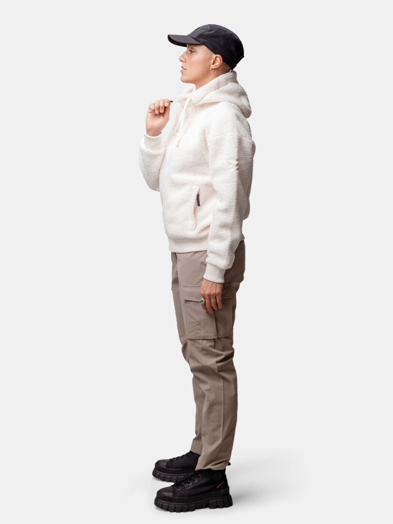 Personen i profil bär Halti Lykky Fleecejacka Dam, beige byxor, svarta skor och svart mössa mot en enfärgad bakgrund.