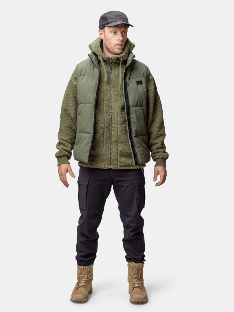 Mannen är klädd i Halti Penger Classic Puffer Väst Herr, olivfärgad fleecejacka, svarta byxor, solbruna stövlar och mörk mössa.